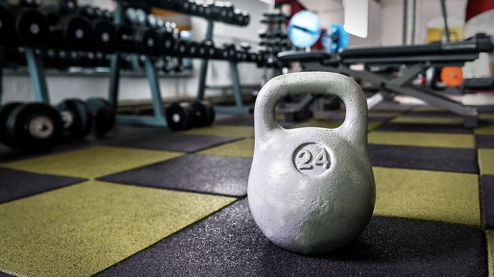 White 24-kettlebell on black-and-yellow gym floor, with blurred dumbbells and bench in the background.