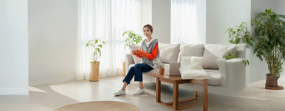 リビングのソファに座り、グレーとオレンジのパーカーとネイビーのジーンズ、白のスニーカーを履いた女性。