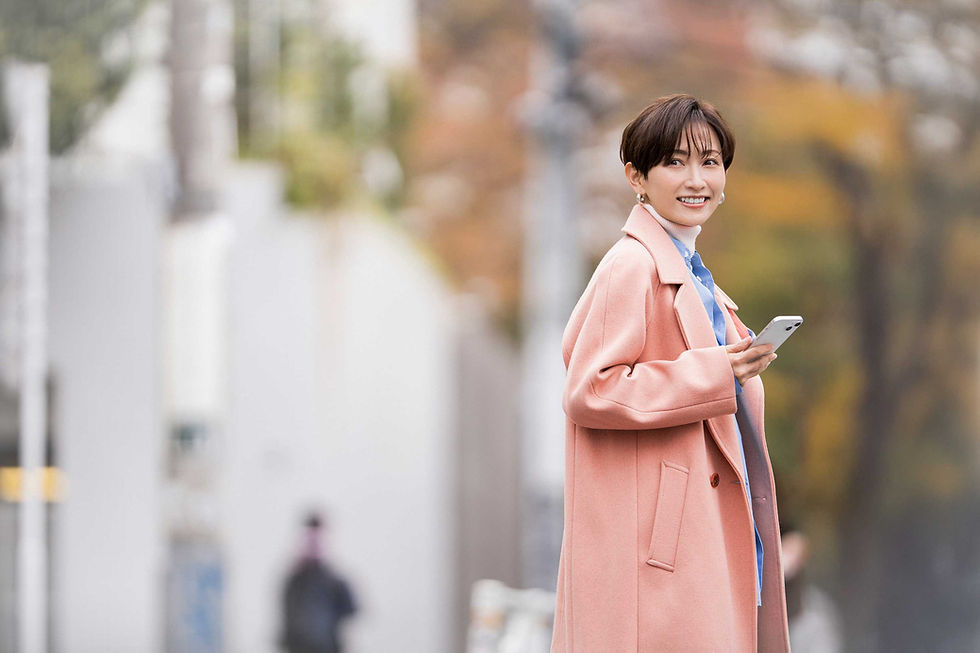  屋外でスマートフォンを持ち、笑顔で振り返るショートヘアの若い女性。ベージュのコートと水色の服を着用している。
