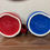 Thumbnail: Pair of Rae Dunn ceramic mugs on wood surface, one red with the phrase 'Star Spangled Cutie' and one blue with 'All American' in white handwritten-style lettering.