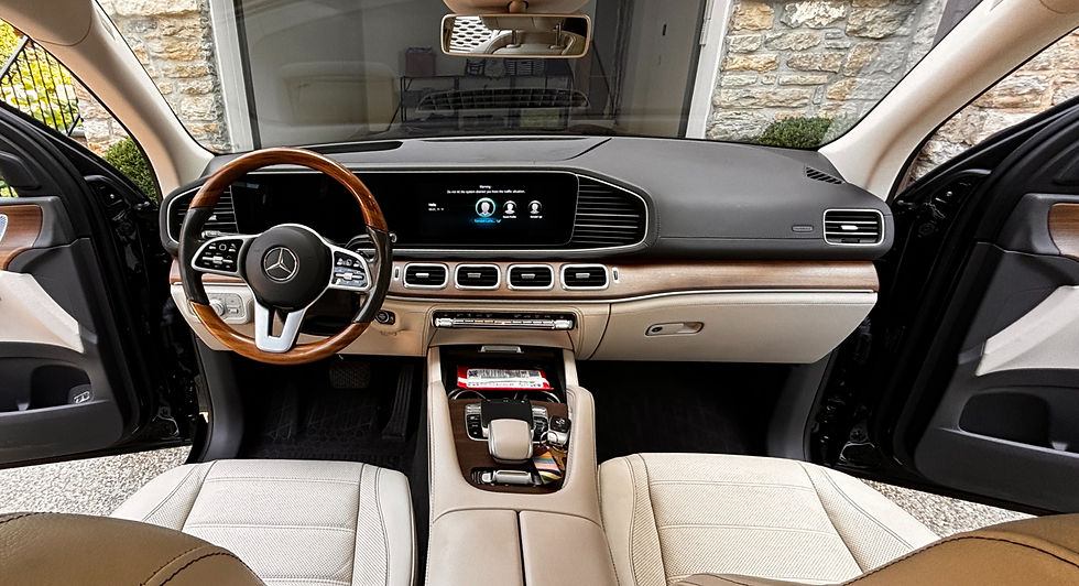 Car interior with cream leather seats, a wooden steering wheel, digital dashboard displaying profiles. Stone wall visible outside.