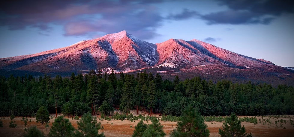 La lumière du crépuscule baigne le paysage, et les majestueux San Francisco Peaks irradient au loin, leurs sommets teintés des roses, des oranges et des ors d’un ciel couchant. Au premier plan, la forêt sombre, paisible, forme un contraste parfait avec l’éclat céleste. Le silence qui émane de cette image est palpable, un moment de grâce où la nature semble s’incliner devant l'immensité du ciel, et où l’horizon révèle la sagesse tranquille des montagnes sacrées.