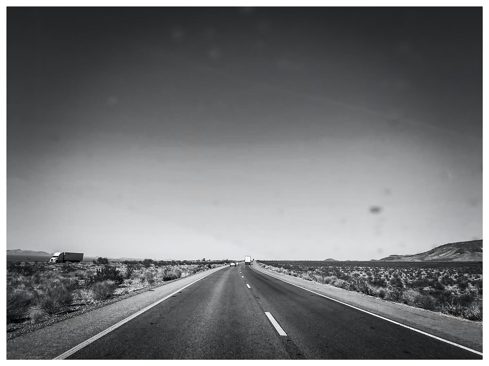 Dans ce noir et blanc intemporel, l'autoroute 40 fend le désert californien, une ligne droite infinie vers l'horizon de l'Arizona. Le bitume, sous le soleil implacable, se perd dans l'immensité poussiéreuse du Mojave. Quelques camions, minuscules points en mouvement, sont les seuls signes de vie sur cette étendue à perte de vue. Le silence est palpable, pesant, comme l'écho d'un monde qui n'appartient qu'à la route. C'est une image de liberté, de solitude et d'un voyage vers l'inconnu, où chaque kilomètre semble murmurer une promesse.