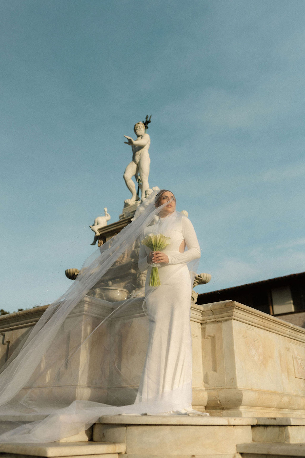 bride with long sleeved dress and cathedral length veil standing in front of marble fountain in amalfi coast elopement