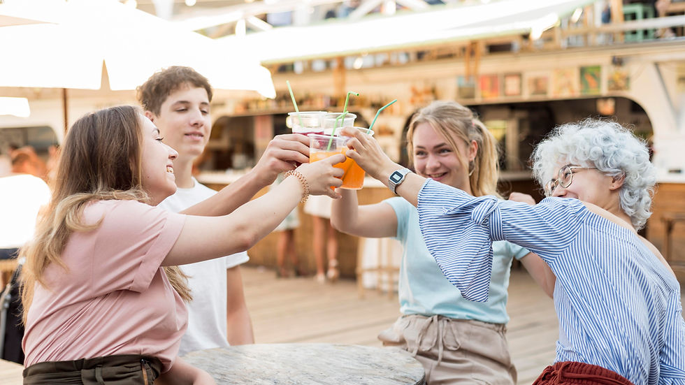 Four friends clink colorful iced drinks at an outdoor café, smiling and laughing in a bright, relaxed scene.