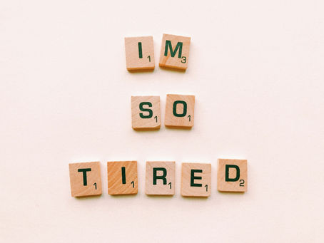 Wooden Scrabble tiles spell "IM SO TIRED" on a light background, conveying exhaustion. Tiles have black letters and point values.