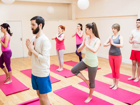 Yoga class of seven adults in tree pose on pink mats in a bright studio with hanging white lanterns, calm and focused.