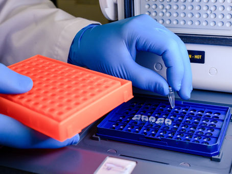Gloved hands place a clear lab tube into a blue rack beside an orange rack and a machine labeled N-HOT in a sterile lab.