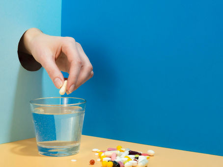 Hand drops a pill into a glass of water beside a pile of colorful pills on a blue tabletop and backdrop.