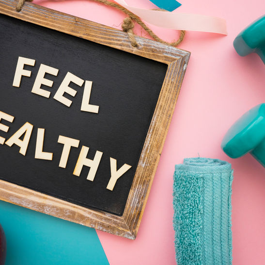 Wooden sign reading FEEL HEALTHY beside teal dumbbells, towel, and apple on a pink-and-turquoise fitness flat lay