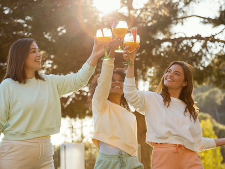 Three women in pastel sweats smile and toast orange drinks outdoors at sunset, with sunlit trees in the background.