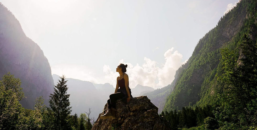 Woman sits in nature on a rock in a sunlit mountain valley, listening to the wisdom of the Earth