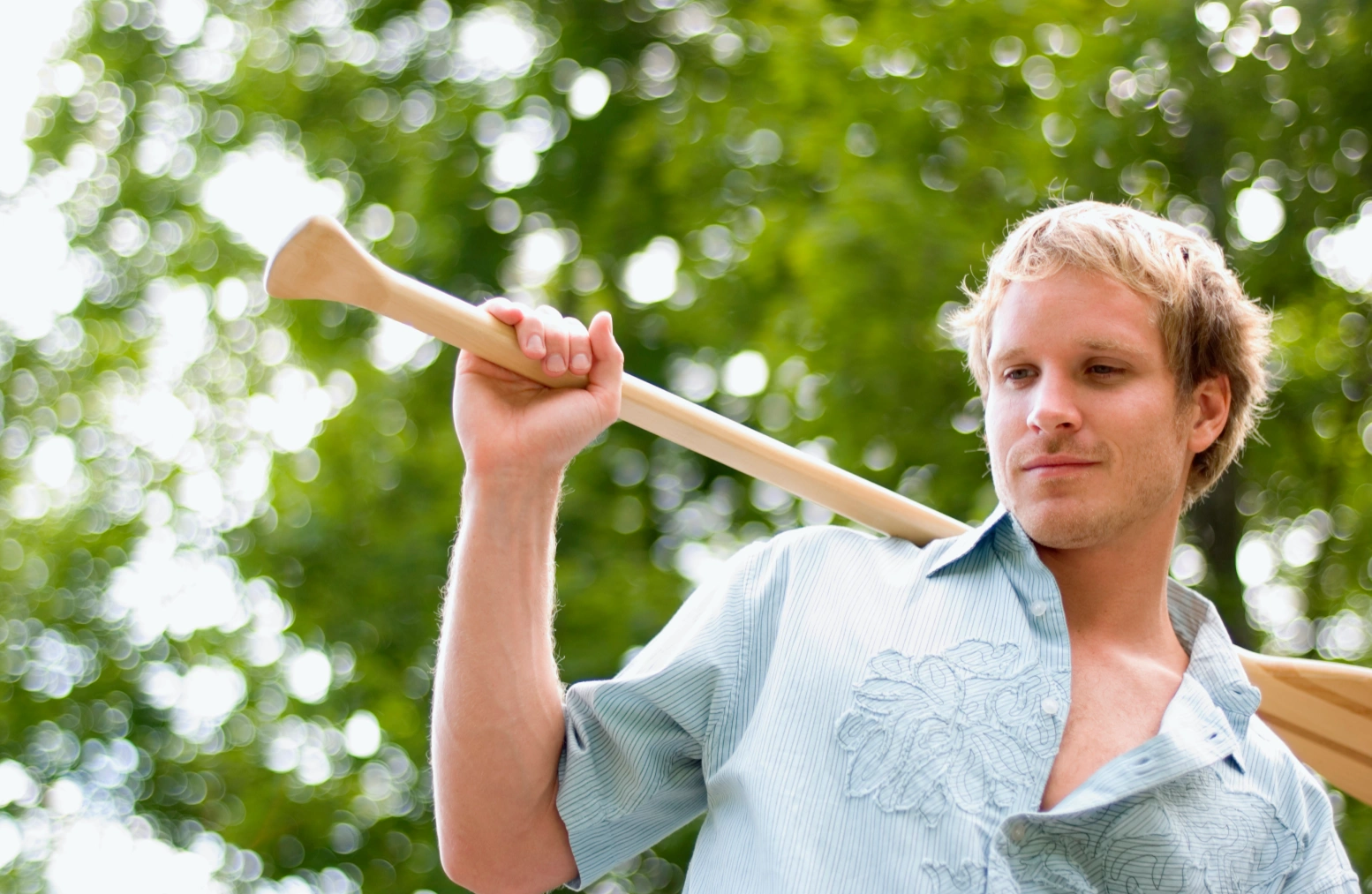 A young man with blonde hair holds a wooden paddle over his shoulder, standing outdoors among blurred greenery. 