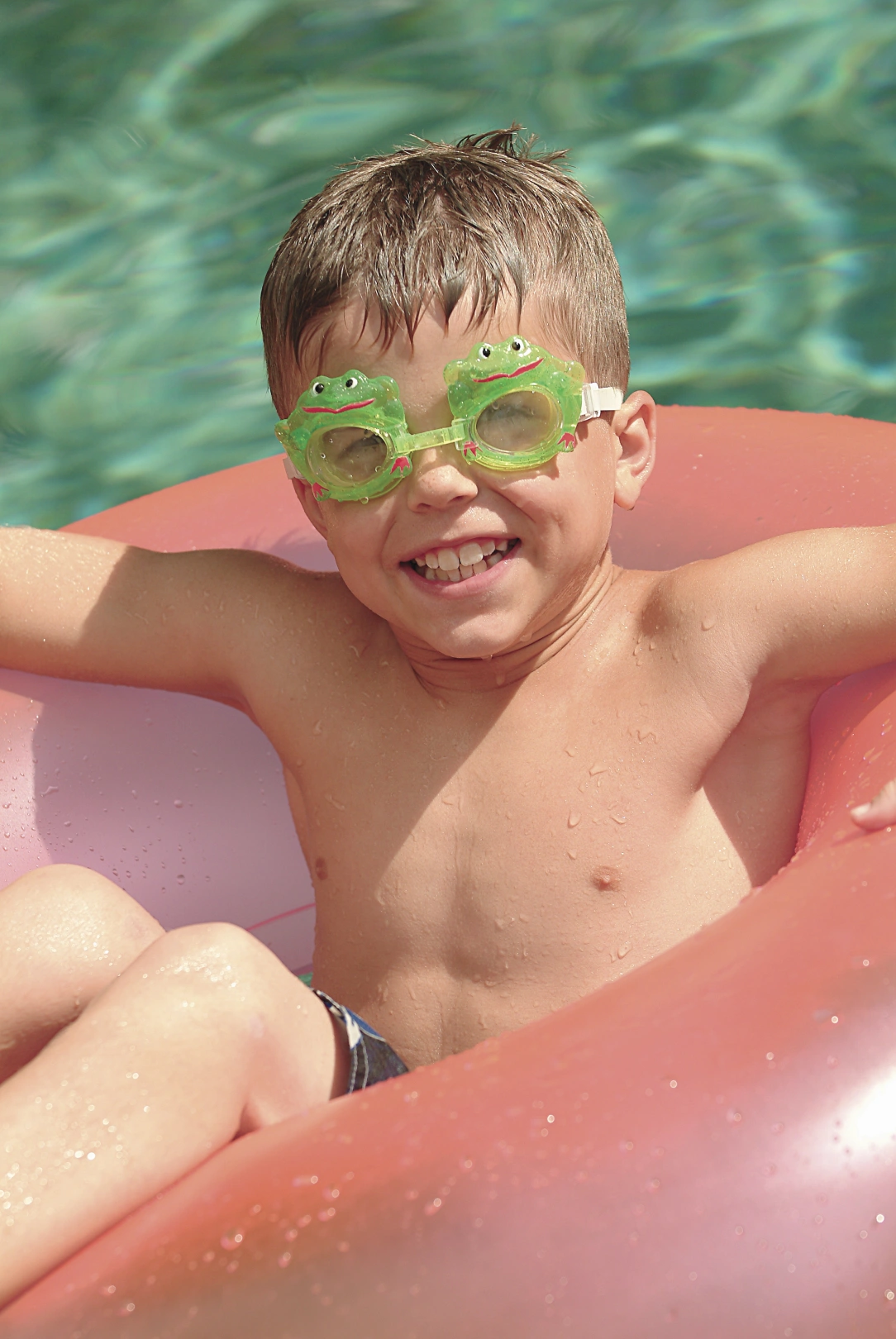 A young boy with short brown hair wears frog-themed goggles while relaxing in a pink inflatable float in a swimming pool. 
