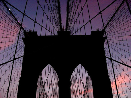Silhouette of a large bridge structure against a colorful twilight sky, featuring intricate cable patterns. 