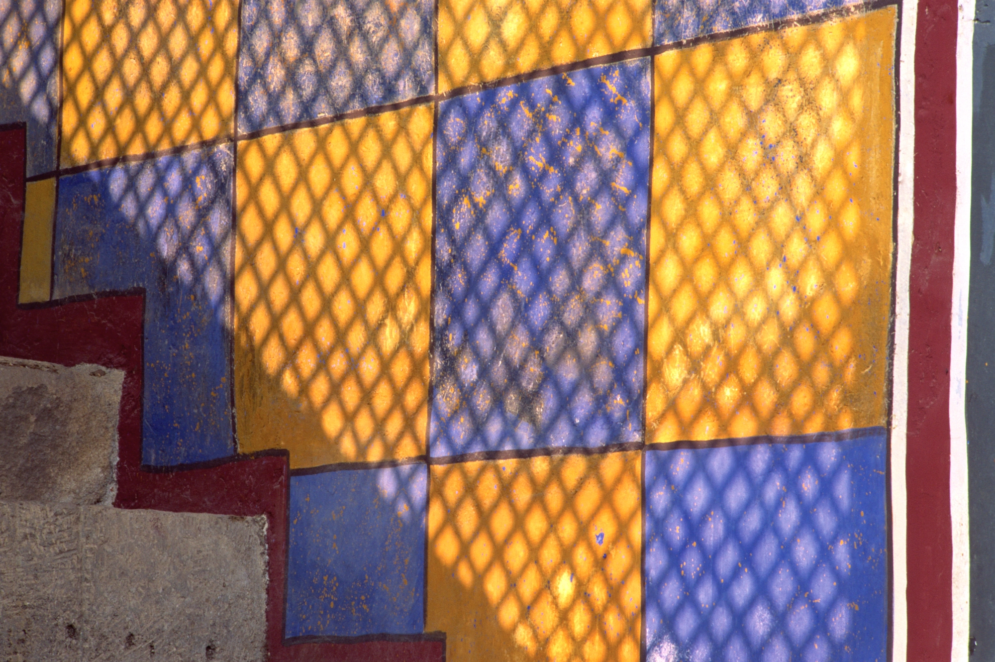 A colorful tiled floor with yellow and purple squares, illuminated by patterned shadows from a lattice structure.
