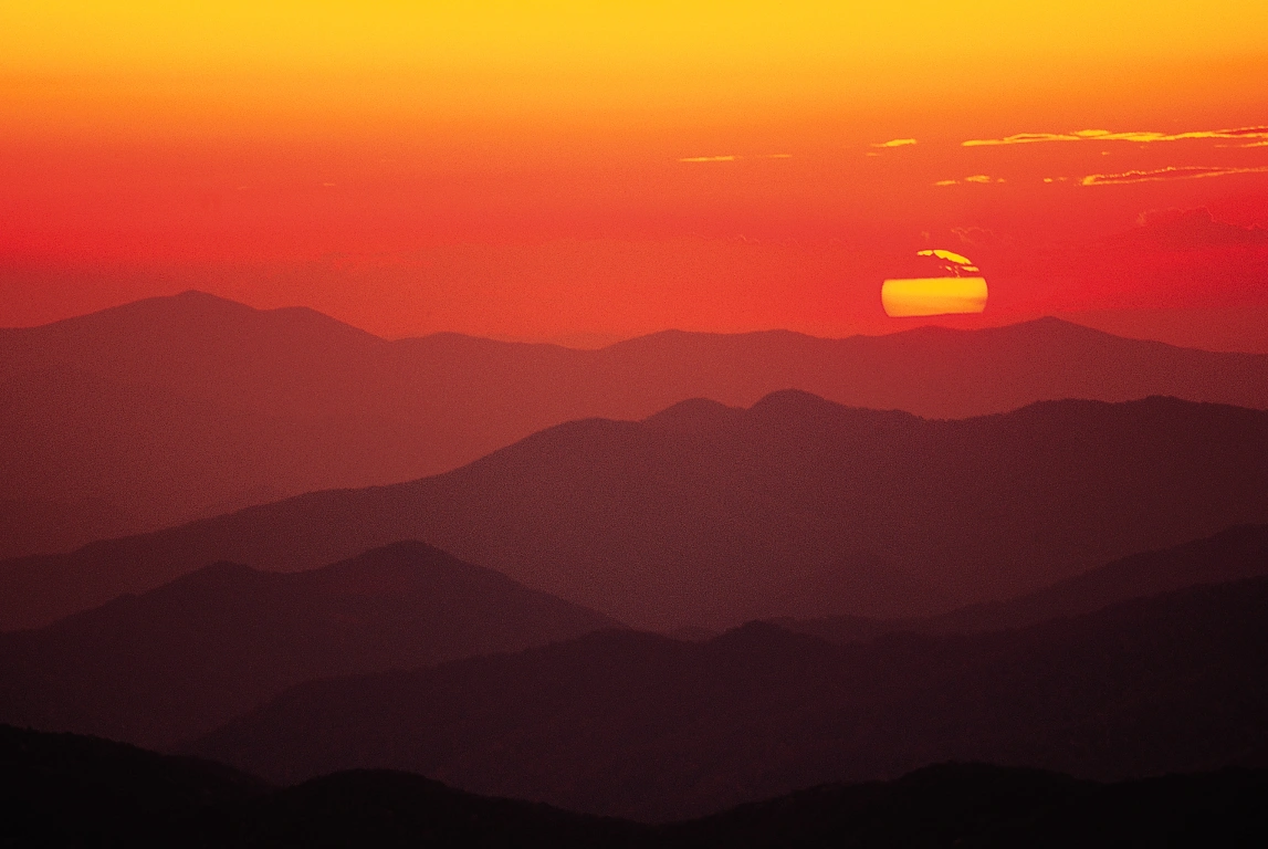 A vibrant sunset casts orange and yellow hues across layered mountain ranges, creating a tranquil atmosphere. 
