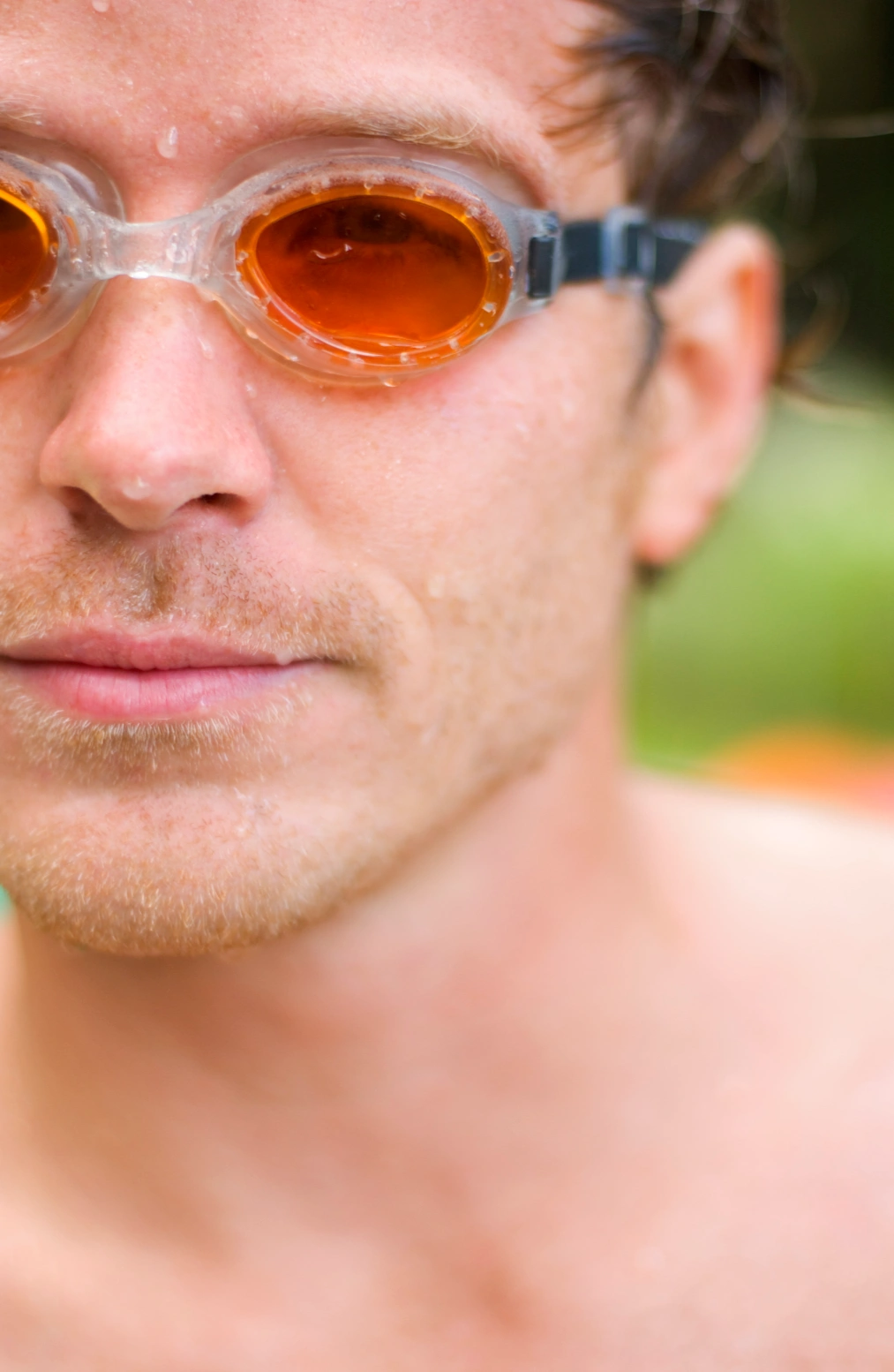 A close-up of a male swimmer wearing orange-tinted goggles, with water droplets on his face. 
