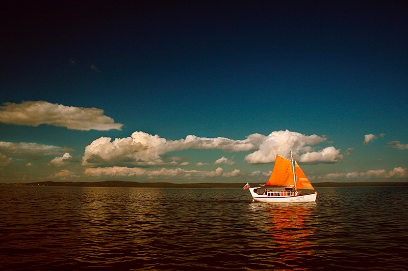 A sailboat with an orange sail glides on calm waters under a vast sky dotted with clouds.