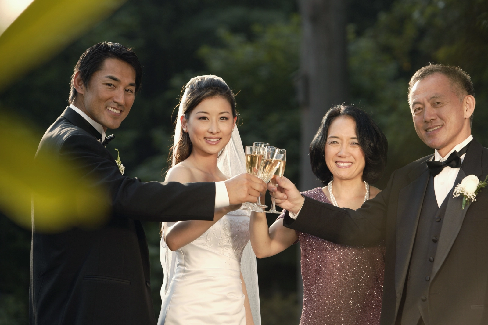 A group of four individuals raise champagne glasses in a toast, dressed in formal attire against a blurred outdoor background. 