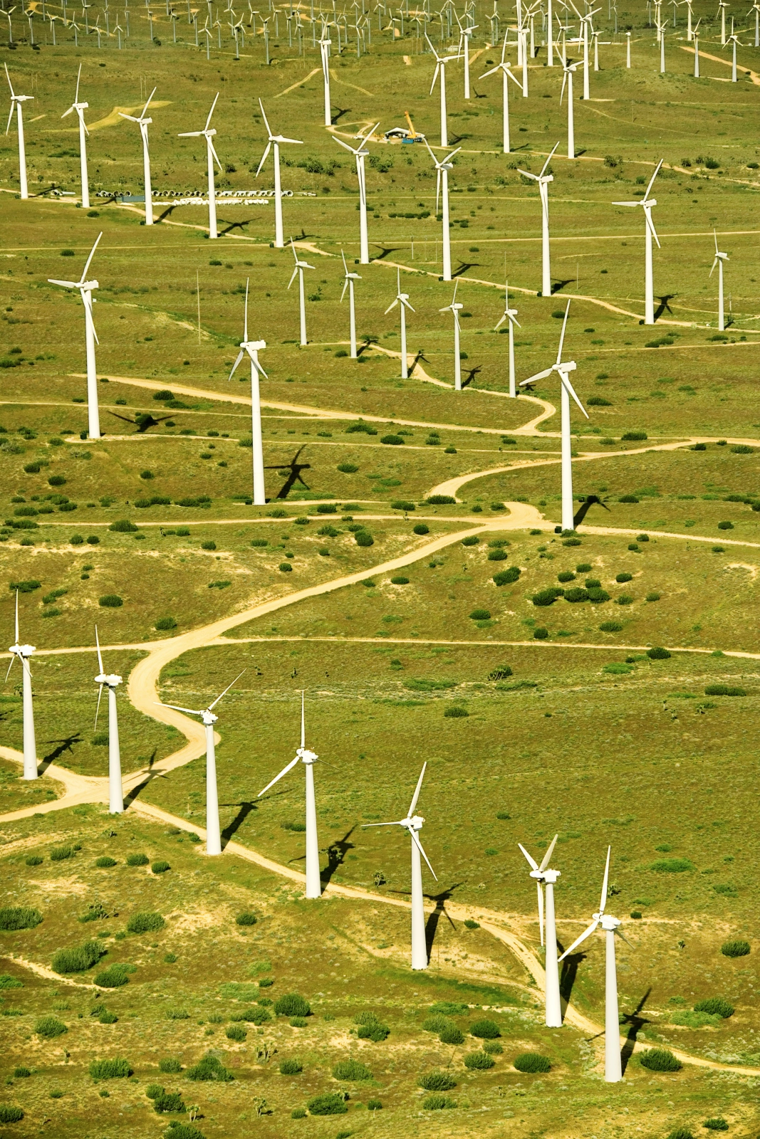Aerial view of a wind farm with numerous wind turbines on rolling green hills, interconnected by winding dirt paths.