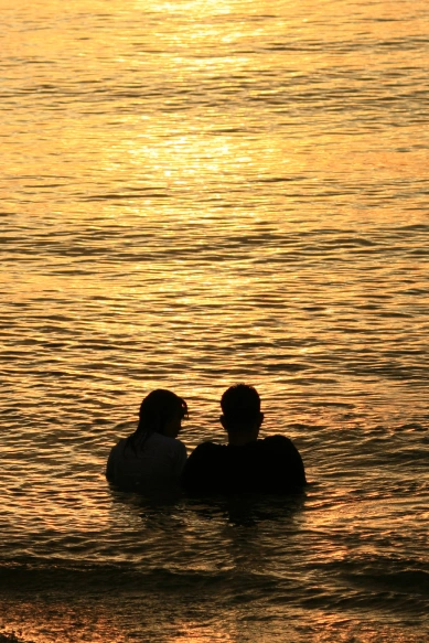 Two figures sit close together in shallow water, silhouetted against a golden sunset reflecting on the surface. 