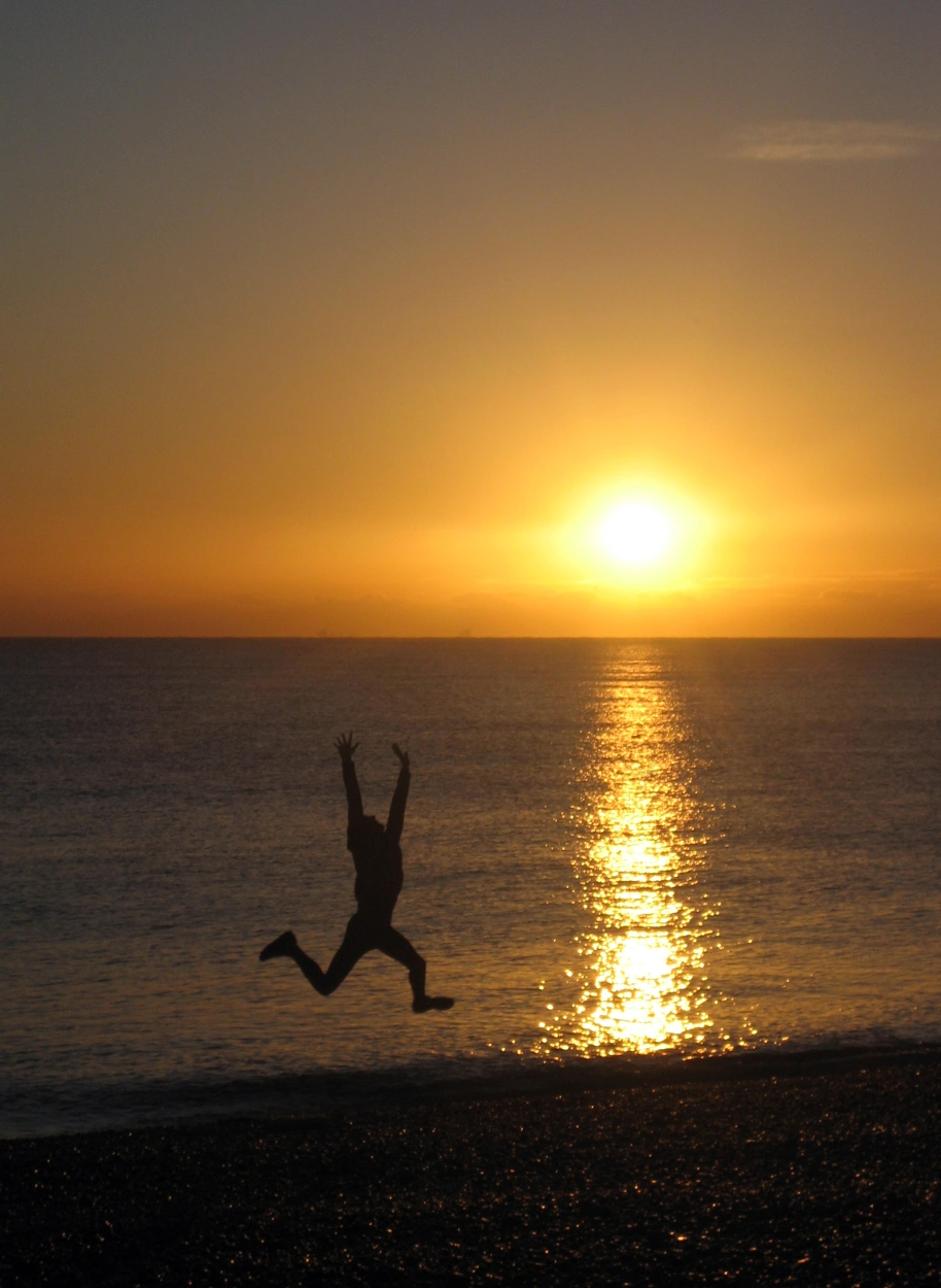 A silhouette of a person jumping near a shimmering ocean at sunset, with warm orange and yellow hues dominating the sky.