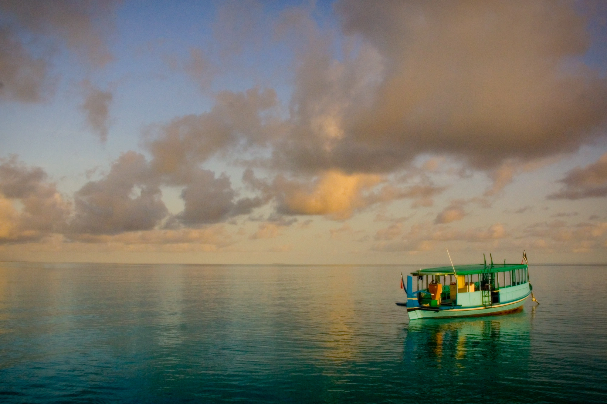 A small boat floats on calm waters under a pastel-colored sky, with soft clouds reflecting in the water. 