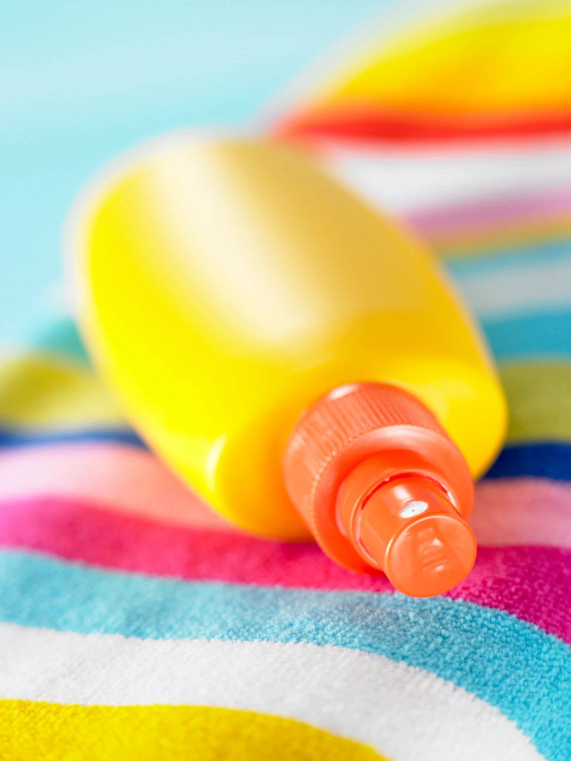 A bright yellow sunscreen bottle with an orange cap is positioned on a striped beach towel featuring vibrant colors. 