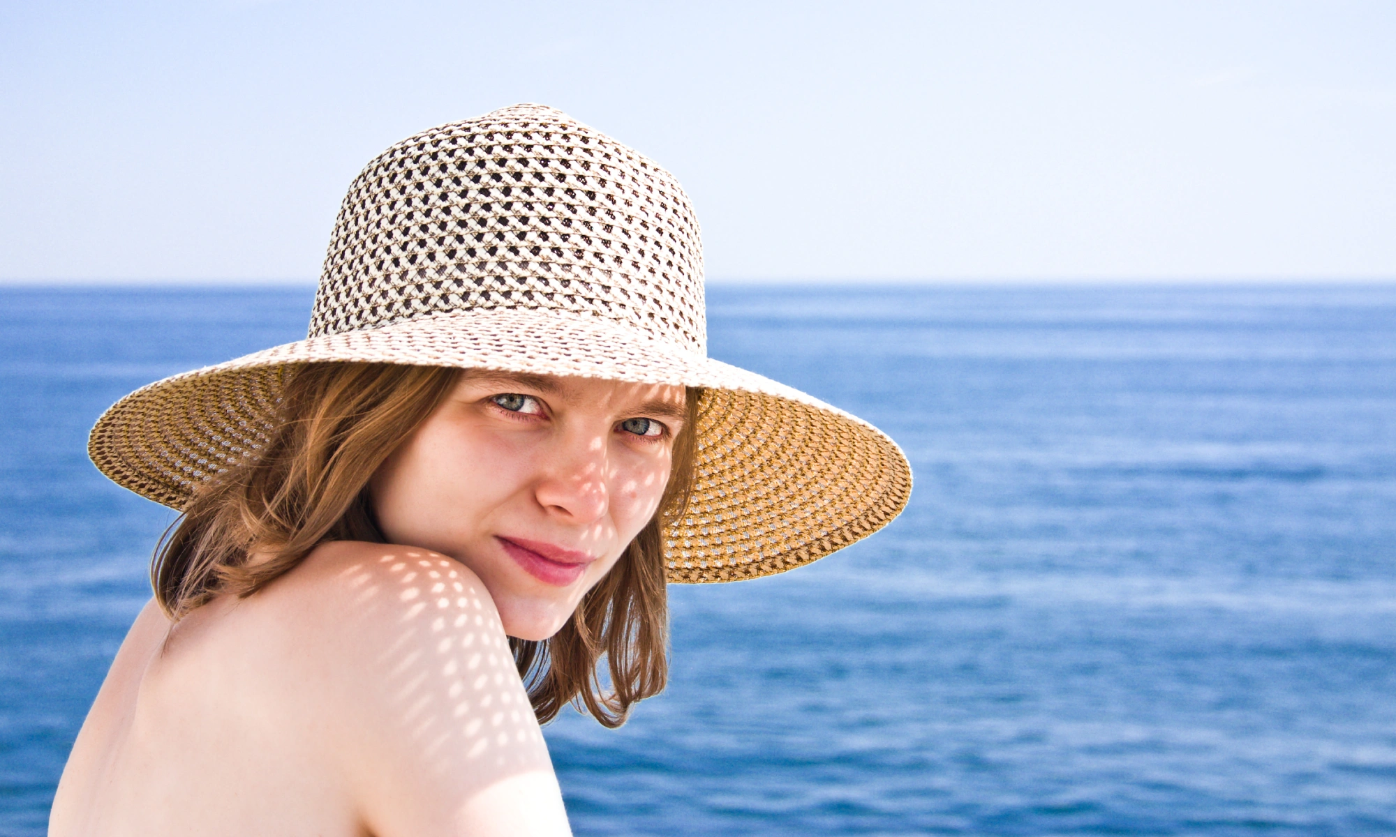 A woman with long hair wears a wide-brimmed straw hat, sitting near calm blue ocean waters.