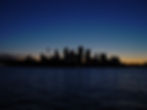 Silhouette of a city skyline against a twilight sky, with water in the foreground reflecting faint lights.