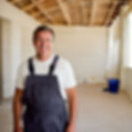 A full-body image of a janitor smiling at the camera in a recently completed and empty room. The space is clean and bright, with a few signs that construction just finished, such as a corner with some paint buckets or traces of building materials left behind.