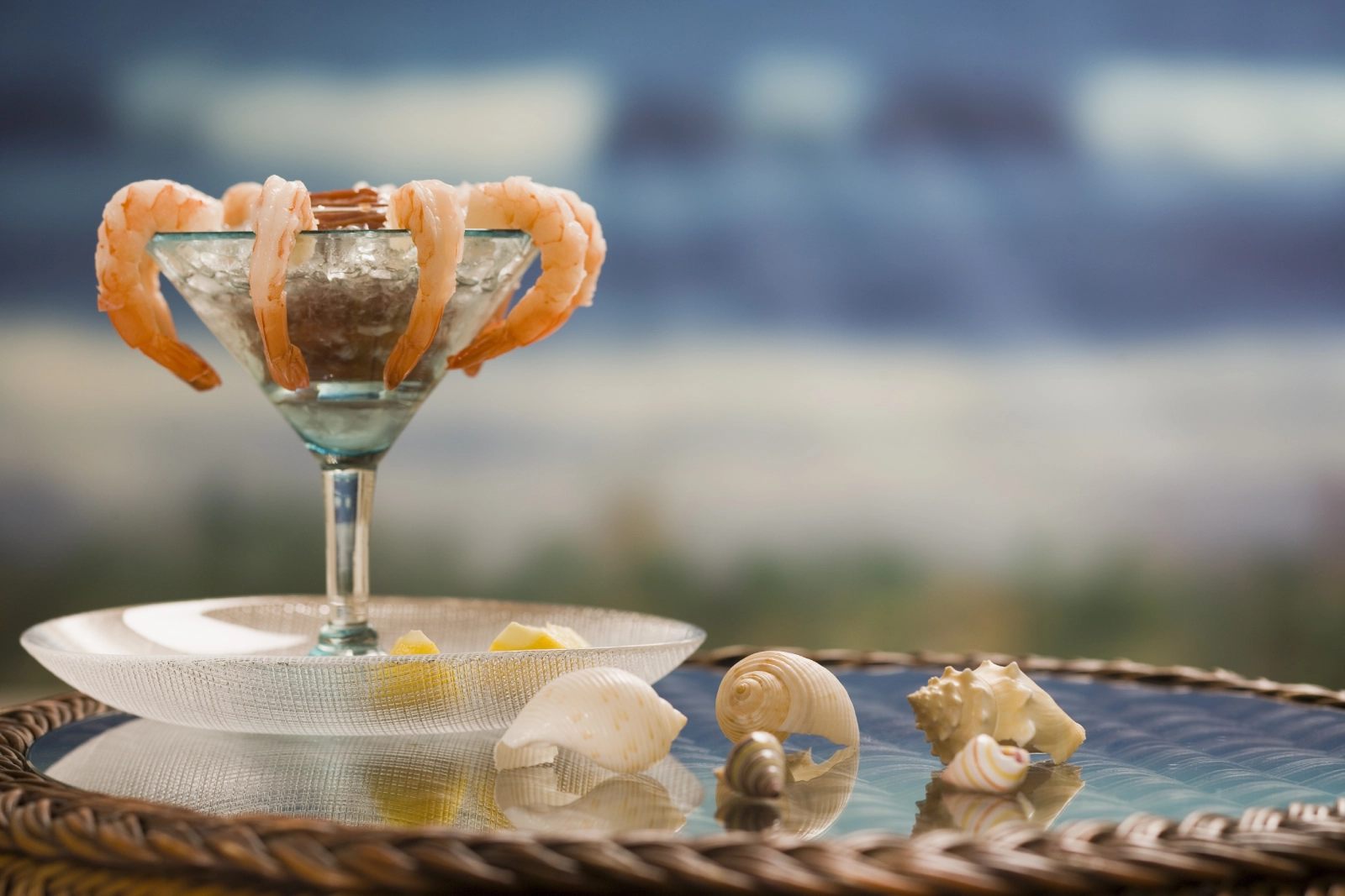A glass martini dish filled with ice holds shrimp garnished around the rim, set on a woven platter.
