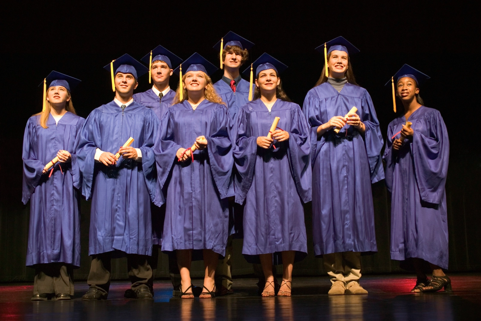 Eight graduates in blue gowns and caps stand together on stage, each holding diplomas, with a dark backdrop.