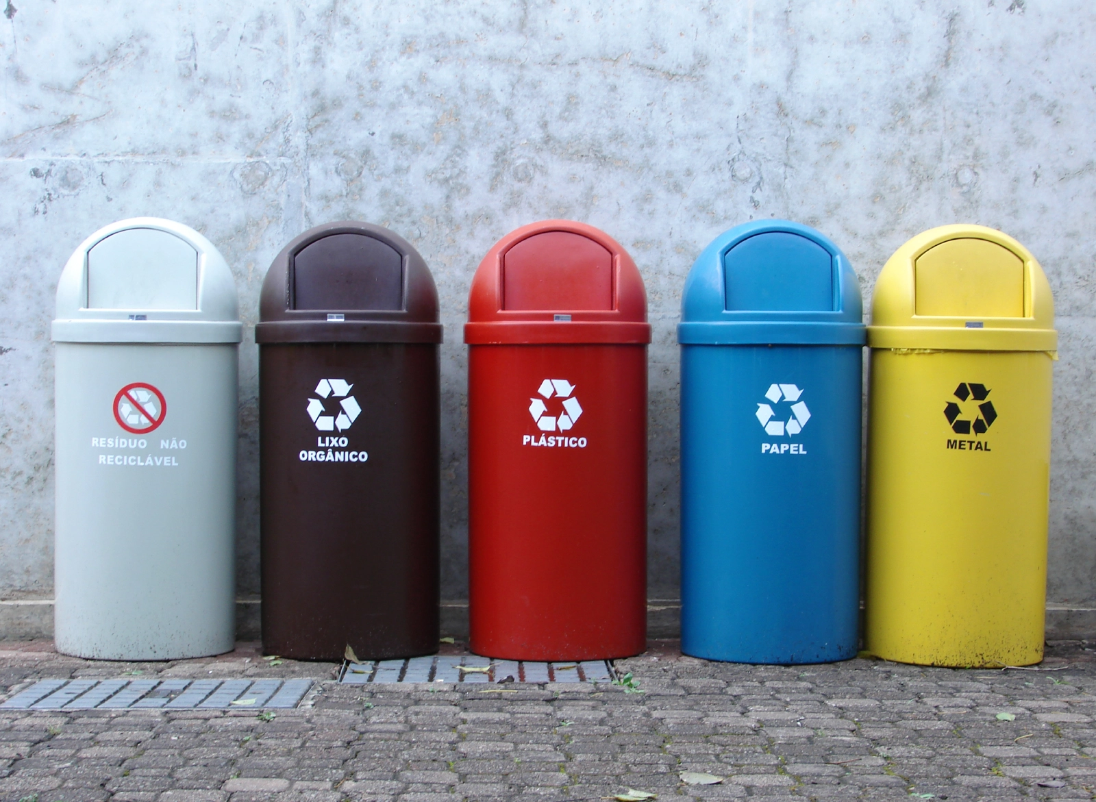 Five recycling bins in various colors—light gray, brown, red, blue, and yellow, arranged against a stone wall