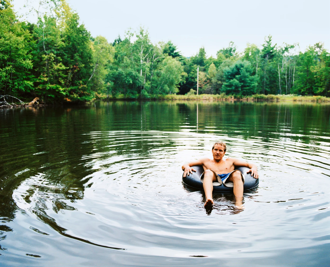 A person floats on a black inner tube in a calm lake surrounded by green trees. 