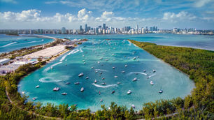 Boats dot bright teal waters in Biscayne Bay, Florida with the Miami skyline in the distance. Lush greenery frames the scene above.