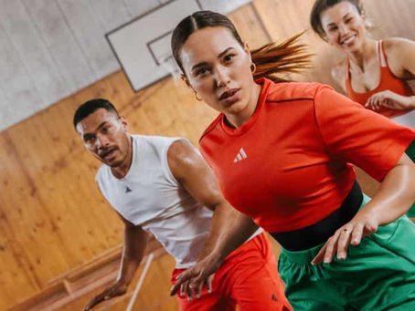 Three people in athletic wear jogging indoors on a wooden floor, focused expressions. Bright colors: red, green, white. Basketball hoop in background.
