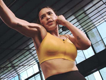 A person in a yellow sports bra, mid-punch, looks focused in a glass-walled gym. Sunlight streams in, highlighting their tattooed arm.