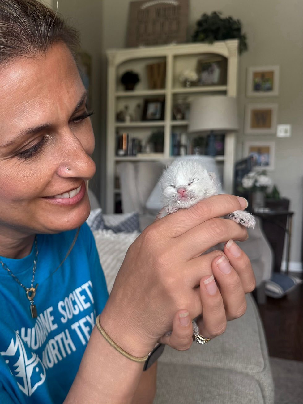 Tiffany, an HSNT foster holds a tiny white kitten in a cozy living room; shirt shows partial North Texas text.