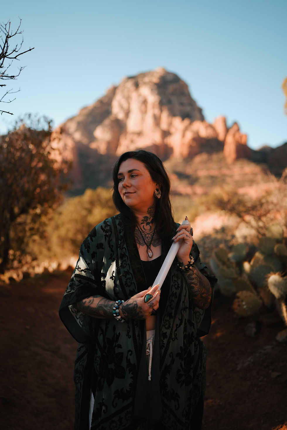 Woman holding a crystal wand in the Sedona desert landscape, wearing patterned clothing and jewelry. Thunder Mountain and cacti in the background. Relaxed mood.