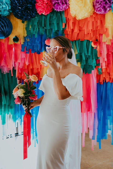 Bride takes a sip of champagne in front of a colourful backdrop.