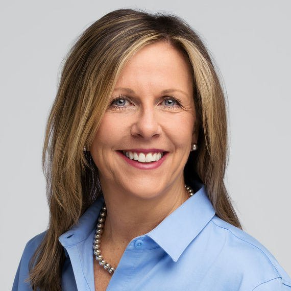 Professional headshot of a woman with light brown hair wearing a blue blouse, smiling against a light gray background