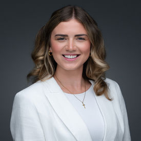Professional headshot of a woman with shoulder length hair wearing a white blazer, smiling against a dark studio background