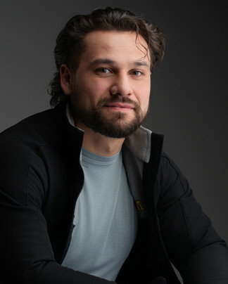 Actor headshot of a man with dark hair and beard wearing a black jacket over a green shirt, looking confidently at camera against a dark background