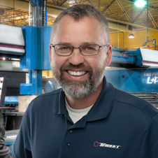 Professional headshot of a man wearing glasses and a polo shirt, smiling in an industrial environment