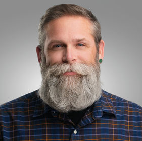Professional headshot of a man with a full gray beard wearing a plaid shirt, neutral studio background
