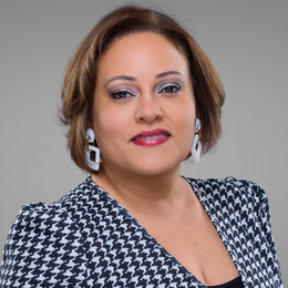Professional headshot of a woman with short hair wearing a patterned jacket and statement earrings, smiling against a neutral background