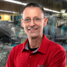 Professional headshot of a man in a red polo shirt, smiling in an industrial workplace setting