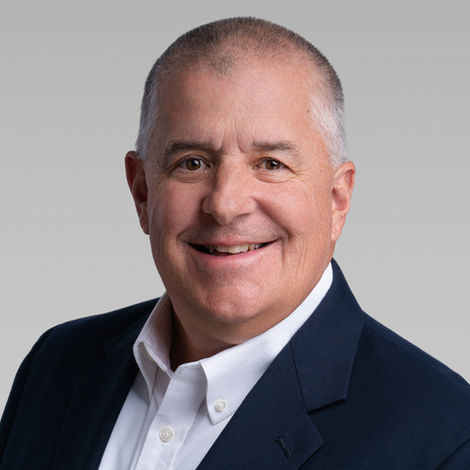 Professional headshot of a middle aged man in a blazer, smiling against a gray studio background